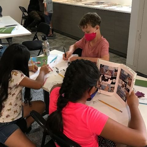 Photo of 3 children, wearing face masks, sitting at a table reading comics and drawing
